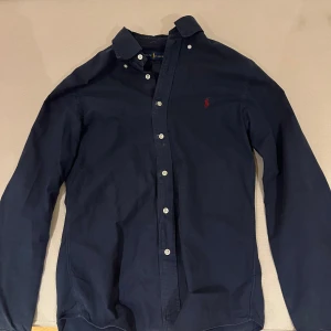 Mörkblå skjorta från Ralph Lauren - Snygg mörkblå skjorta från Ralph Lauren i slim fit. Skjortan har långa ärmar och knappar framtill. Den är broderad med det klassiska röda logotypen på bröstet. Perfekt för en stilren look. Varsamt använd ! 