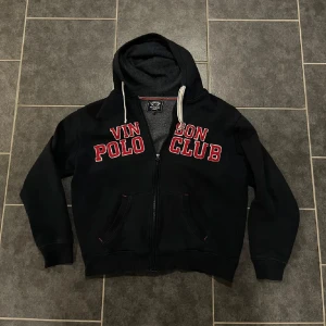 Mörk blå hoodie från Vinson Polo Club - Säljer en svart hoodie från Vinson Polo Club med röd text på bröstet. Den har en dragkedja framtill och en mysig huva med snören. Perfekt för kyliga dagar. Jag är 185 och den är lite kort på mig. Runt 3 cm. Så om du är 180 skulle den nog sitta bra