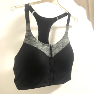 Svart sport-bh med dragkedja - Snygg och funktionell svart sport-bh med grå detaljer och dragkedja framtill. Perfekt för träning med justerbara axelband och brottarrygg för extra stöd.