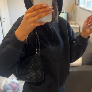 Svart glittrig axelväska - Snygg svart axelväska med glittriga detaljer. Perfekt för att ge din outfit lite extra bling. Väskan har en praktisk storlek och en justerbar axelrem.