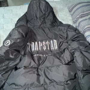 Svart dunjacka från Trapstar - Säljer en svart dunjacka från Trapstar med huva och dragkedja. Jackan har tryck på ryggen och en logotyp på ärmen. Perfekt för kalla dagar, den är aldrig använd och passar Xs och S, priset kan diskuteras, betala tryggt på plick appen och extra bilder kan alltid skickas!