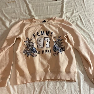 Ljus rosa tröja från Gina Tricot - Säljer en ljus rosa tröja från Gina Tricot med texten 'FEMME 97 AUTH. EST.' och blommiga broderier i blått och rosa. Tröjan har långa ärmar och ribbade muddar vid ärmslut och nederkant. Perfekt för en avslappnad stil! Säljer då den inte används. OBS tröjan har lite fläckar ovanför texten.
