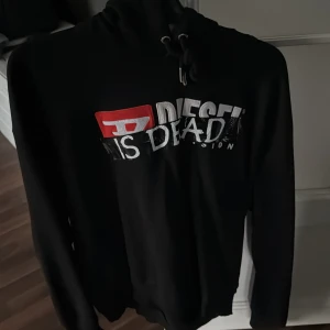 Svart hoodie från Diesel - Säljer en svart hoodie från Diesel med tryck 'Diesel is Dead' i vitt och rött på framsidan. Tröjan har en klassisk huva med dragsko och långa ärmar. Perfekt för en avslappnad stil.