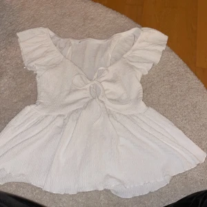 Vit blus med knytning - Charmig vit blus med korta ärmar och knytning framtill. Blusen har en volangdetalj vid ärmarna och en lätt rynkad struktur som ger en romantisk känsla. Har klippt bort lappen men den är från lager 157!