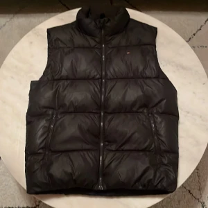 Svart dunväst från Tommy Hilfiger - Snygg svart dunväst från Tommy Hilfiger med dragkedja och hög krage. Västen har märkets logga på bröstet och ett stort tryck på ryggen. Perfekt för kyliga dagar. Väldigt bra skick och knappt använd! OBS frakt börjar på 68kkr