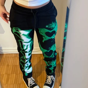 Svarta mjukisbyxor med grönt mönster från Jaded LDN - Coola svarta mjukisbyxor med grönt graffiti-liknande mönster och hjärtan. Byxorna har en elastisk midja med snörning för justerbar passform. Perfekta till y2k stil.