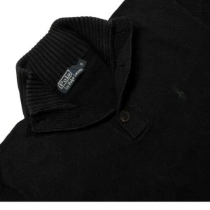 Svart tröja från Polo Ralph Lauren - Snygg svart tröja från Polo Ralph Lauren med ribbad krage och knappdetalj. Den har ett diskret broderat logotypmärke i grönt på bröstet. Perfekt för en stilren look.                                                   Ralph Lauren Tröja Mycket fin skick  Storlek: S Längd: 65cm Bredd: 53 cm x 2
