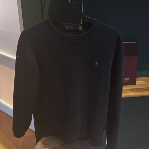 Svart tröja från Polo Ralph Lauren - Säljer en stilren blå tröja från Polo Ralph Lauren med det klassiska röda logotypen på bröstet. Tröjan har en rund halsringning och långa ärmar, perfekt för en avslappnad look samt perfekt nu när vädret blir mer kyligt 