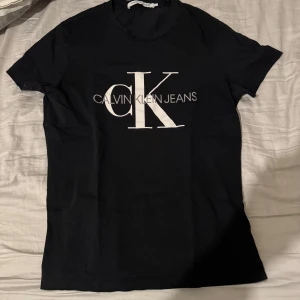 Svart t-shirt från Calvin Klein Jeans - Snygg svart t-shirt från Calvin Klein Jeans med stort vitt CK-logotryck på framsidan. Perfekt för en stilren och avslappnad look. Kortärmad och tillverkad i mjukt material för extra komfort. T-shirten är storlek L, men jag kommer att lägga storlek M eftersom den passar bäst på storlek M