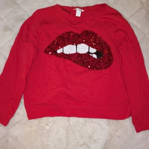 Röd tröja med paljettläppar från H&M - Säljer en cool röd tröja från H&M med ett stort paljettmotiv av läppar på framsidan. Tröjan har långa ärmar och en avslappnad passform. Perfekt för att ge din outfit en färgklick! 💋