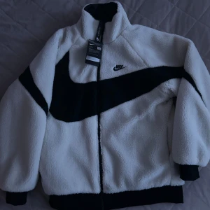 Vit fleecejacka från Nike - På stockx ”nike big swoosh reversible boa jacket”. En HELT NY och snygg vit fleecejacka från Nike med svart swoosh-design. Jackan har dragkedja och hög krage för extra värme. Perfekt för kyliga dagar. Den är storlek EUR S. Dm för mer info.                                                             PRISET KAN DISKUTERAS!!