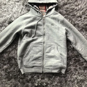 Burberry zip hoodie - Snygg grå huvtröja från Burberry med dragkedja och klassiskt rutigt mönster i huvan. Tröjan har långa ärmar och praktiska fickor framtill. Den är i fint skick och finns inga direkta tecken på slitning. Hör av dig vid frågor och funderingar. Mvh Emil