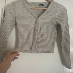 Grå ribbad kofta från Gina Tricot - Säljer en stilren grå ribbad kofta från Gina Tricot i storlek XS. Koftan har en v-ringad design med knappar framtill och långa ärmar. Perfekt för lager-på-lager under kyligare dagar.