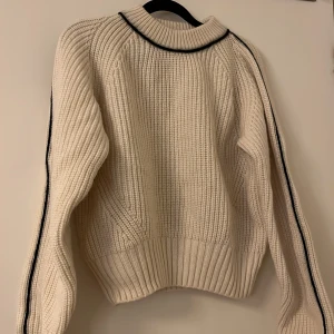 Vit stickad tröja med svarta detaljer - Säljer en stilren vit stickad tröja med svarta detaljer längs kragen och ärmarna. Tröjan har en ribbad struktur och långa ärmar, perfekt för kyligare dagar.