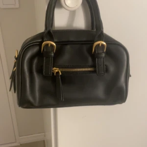 Svart handväska från Zara - Snygg svart handväska från Zara i skinn med guldfärgade detaljer. Väskan har en praktisk dragkedja och två handtag. Insidan är fodrad med ett randigt mönster och har flera fack för enkel organisering, priset kan ändra vi snabb affär☺️🩷