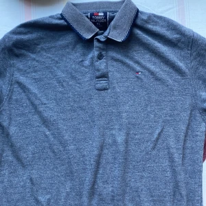Grå pikétröja från Tommy Hilfiger - Snygg grå pikétröja från Tommy Hilfiger i storlek XL. Tröjan har en klassisk krage med knappar och det ikoniska logotypmärket på bröstet. Perfekt för en stilren och avslappnad look.