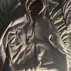 Grå huvtröja från Burberry - Säljer en grå huvtröja från Burberry med klassiskt rutigt mönster i huvan. Tröjan har dragkedja och fickor framtill. Perfekt för en stilren och bekväm look.