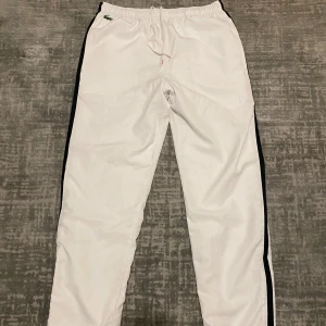 Vita träningsbyxor från Lacoste - Snygga vita träningsbyxor från Lacoste med svarta ränder längs sidorna. Byxorna har en elastisk midja med snörning och dragkedjor vid bensluten för extra komfort. Perfekta för sportiga aktiviteter.