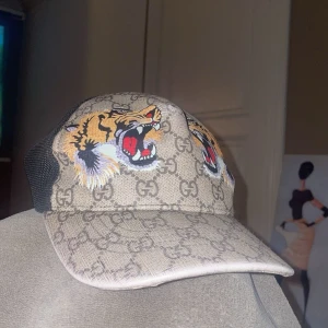 Beige keps med tigertryck - Snygg beige keps från Gucci med ett djärvt tigertryck och det ikoniska GG-mönstret. Kepsen har en justerbar rem med gröna och röda ränder baktill. Perfekt för att ge din outfit en lyxig touch.  Lapparna är bort dragna på grund utav att en gick av så drog jag av dom andra också 