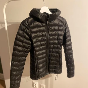 Svart dunjacka från Everest - Säljer en svart dunjacka från Everest med huva och dragkedja. Jackan har en quiltad design och är perfekt för kyligare väder. Den är lätt och smidig att bära med sig.