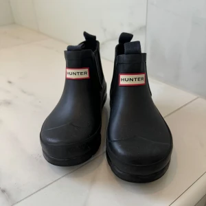 Svarta boots från Hunter - Säljer ett par svarta boots från Hunter i mycket bra skick. De är perfekta för regniga dagar och har en stilren design med Hunter-loggan framtill. Bootsens material är robust och vattentätt, vilket gör dem idealiska för höst och vår. De har en bekväm passform och är lätta att ta på och av.