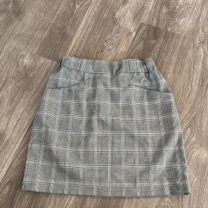Grå rutig kjol - Snygg grå rutig kjol i normal passform. Perfekt för både vardag och festliga tillfällen. Kjolen har en klassisk design med ett stilrent rutmönster som ger en tidlös look. Passar bra till både blusar och t-shirts.
