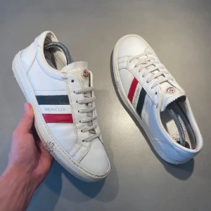 Vita sneakers från Moncler - Snygga vita sneakers från Moncler med klassiska ränder i rött och blått på sidan. Skorna har snörning och är i mycket bra skick. Perfekta för både vardag och festliga tillfällen. Storlek 42 men passar 43