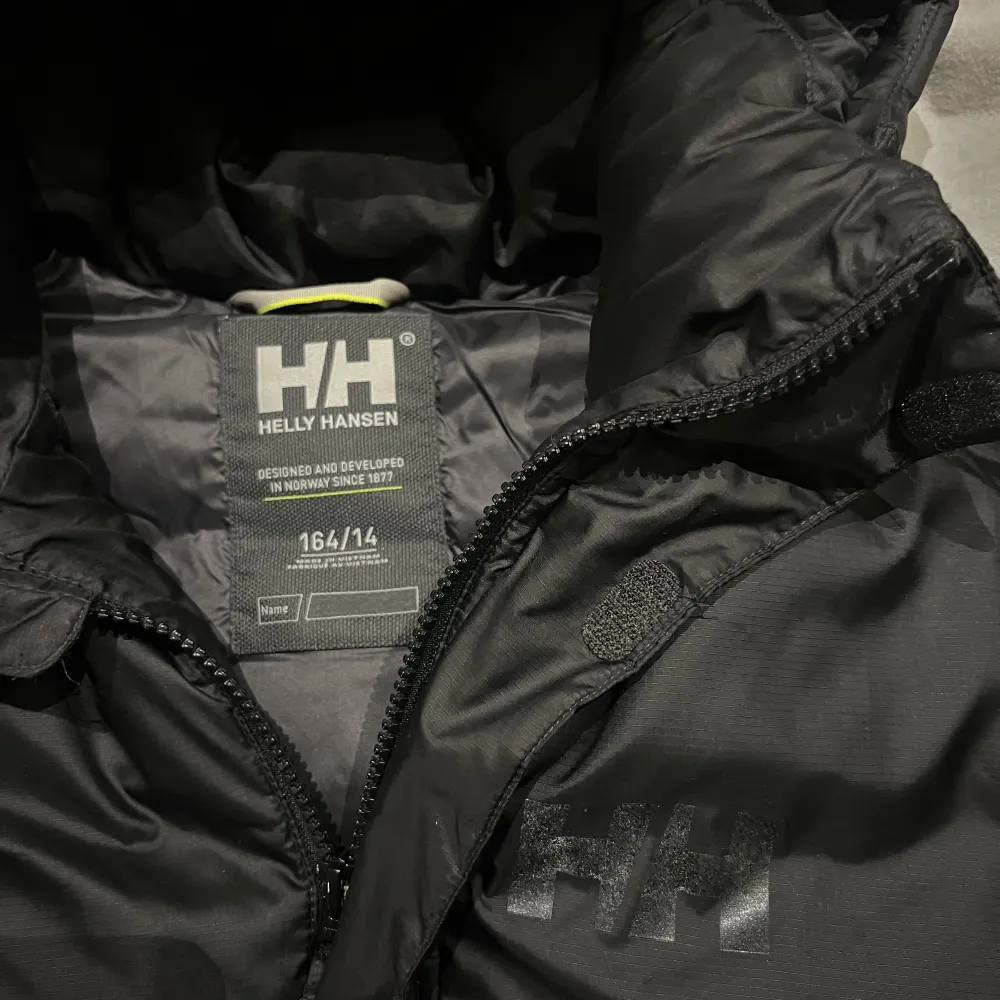 Säljer en svart dunjacka från Helly Hansen i storlek 164. Jackan är perfekt för kalla vinterdagar med sin tjocka vaddering och huva. Den har en stilren design med Helly Hansen-logga på bröstet och ryggen. Jackan är i mycket bra skick och redo för nya äventyr! 🖤Luvan är avtagbar. . Takit.