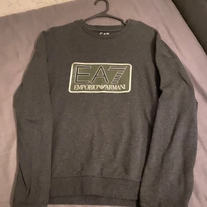 Grå sweatshirt från Emporio Armani - Säljer en snygg grå sweatshirt från Emporio Armani med deras ikoniska EA7-logga på framsidan. Tröjan är i bra skick och perfekt för både vardag och chill. Den har långa ärmar och en bekväm passform. Perfekt för höst och vinter!