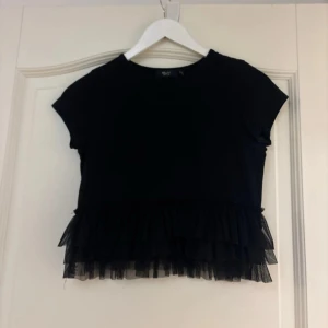 Svart topp med volangdetaljer - Säljer en snygg svart topp med korta ärmar och volangdetaljer i nederkant. Toppen är i mycket bra skick och har en normal passform. Barn storlek men passar även XS