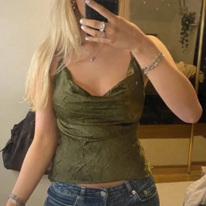 Olivgrön satin topp med smala axelband - Säljer en snygg olivgrön topp med smala axelband och draperad framsida. Toppen är i ett glansigt material som ger en lyxig känsla. Perfekt för en utekväll eller fest! Har litet hål som syns på  sista bilden, skulle inte säga att det syns när man har den på sig så länge man inte drar i den som ja gjorde på bilden 🍂🧸☁️🪐🕯️