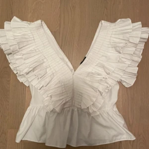 Vit blus med volangdetaljer - Säljer en superfin vit blus med djup v-ringning och volangdetaljer längs ärmarna. Perfekt för sommarens fester eller en stilren vardagslook. Blusen har en luftig passform och är tillverkad i ett lätt material som känns skönt mot huden. Finns en defekt att öglan på toppen har gått sönder men knappen är kvar! Enkel i fixa med en tråd. 