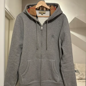 Grå hoodie från Burberry - Säljer en snygg grå hoodie från Burberry i storlek L. Den har en klassisk dragkedja och en mysig huva med det ikoniska Burberry-mönstret på insidan. Perfekt för höst och vinter, och passar till både vardag och chill. 🧥