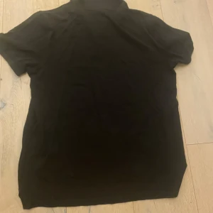 Svart pikétröja från Hugo Boss - Säljer en stilren svart pikétröja från Hugo Boss i bra skick. Tröjan har korta ärmar och en klassisk krage med knappar. Perfekt för både vardag och festliga tillfällen. Passar bra till jeans eller chinos för en avslappnad men ändå uppklädd look.