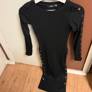 Svart ribbad klänning från CBR - Snygg svart ribbad klänning från CBR i storlek S. Den har långa ärmar och knappar längs sidan och ärmen för en cool detalj. Perfekt för höst och vinter, både till vardags och fest. Klänningen är tight och framhäver figuren på ett stilrent sätt.