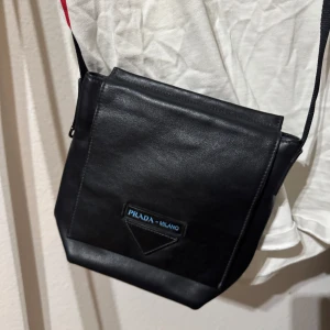 Svart axelväska från Prada - Snygg svart axelväska från Prada i skinn. Perfekt för både vardag och fest. Väskan har en enkel design med en justerbar axelrem och en diskret logga på framsidan. Den är i mycket bra skick och passar till alla outfits. En klassisk och tidlös accessoar för alla tillfällen.