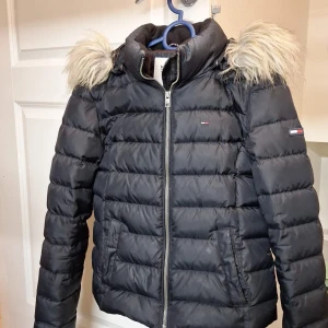 Svart dunjacka från Hilfiger Denim - Säljer en snygg svart dunjacka från Hilfiger Denim med pälskantad huva. Jackan har en dragkedja framtill och är perfekt för kalla vinterdagar. Den är i bra skick och har en klassisk design med långa ärmar och quiltad stil. Perfekt för både vardag och fest!