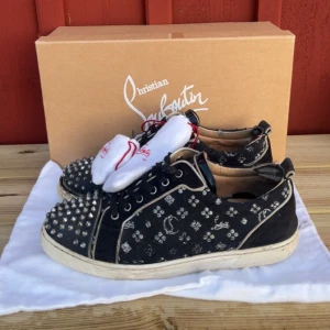 Svarta sneakers med nitar från Christian Louboutin - Säljer ett par coola svarta sneakers från Christian Louboutin med nitar på tån och ett mönster av vita symboler. Skorna har den klassiska röda sulan och är i bra skick. Perfekta för att ge din outfit en edgy touch! Storlek 45 