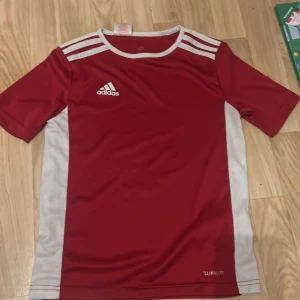 Röd och vit fotbollströja från Adidas - Säljer en snygg röd och vit fotbollströja från Adidas i Climalite-material. Perfekt för träning eller match! Tröjan har korta ärmar och de klassiska tre ränderna på axlarna. Den är i mycket bra skick och redo för nya äventyr på fotbollsplanen. Passar perfekt för unga spelare!