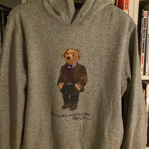 Grå hoodie från Ralph Lauren med nallemotiv - Säljer en superfin grå hoodie från Ralph Lauren med ett gulligt nallemotiv på framsidan. Perfekt för en avslappnad stil och passar bra till både jeans och joggers. Hoodien är i bra skick och har en skön passform. Perfekt för höst och vår!