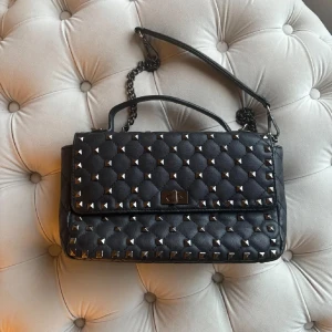 Svart quiltad axelväska med nitar - Säljer en supercool svart quiltad axelväska med nitar och kedjehandtag som är ”valentino” liknande. Perfekt för att ge din outfit en edgy touch. Väskan har en praktisk storlek och stängs med en snygg låsdetalj framtill. Passar både till vardags och fest! D