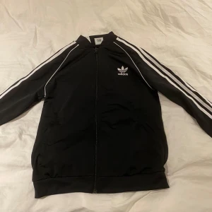 Svart träningskofta från Adidas - Säljer en svart träningskofta från Adidas i nyskick. Den har de klassiska vita ränderna längs ärmarna och en dragkedja framtill. Passar alla årstider.