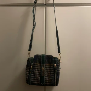 Axelväska - Säljer en superfin rutig axelväska med gröna detaljer och dragkedja. Väskan har ett klassiskt rutmönster i olika färger och en justerbar axelrem. Perfekt för höst och vinter. Den här även ett till handtag. Helt oanvänd.