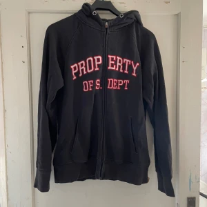 Svart hoodie med tryck - Säljer en svart hoodie med texten 'PROPERTY OF S. DEPT' i rött och vitt på framsidan. Den har en dragkedja och en bekväm huva. Perfekt för en avslappnad stil eller kyliga dagar. Passar bra till jeans eller mjukisbyxor. 🖤
