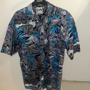 Färgglad kortärmad skjorta från T&S Company - Säljer en cool och färgglad kortärmad skjorta från T&S Company. Skjortan har ett unikt mönster i blått, grått och svart med inslag av lila. Perfekt för sommarens fester eller en avslappnad dag på stan. Den är i nyskick och redo för nya äventyr!