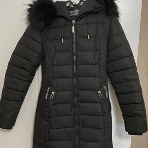 Svart dunjacka från Garoff - Säljer en snygg svart dunjacka från Garoff i storlek L. Jackan har en fluffig pälskantad huva och dragkedja framtill. Perfekt för kalla vinterdagar med sin långa design och värmande material. 