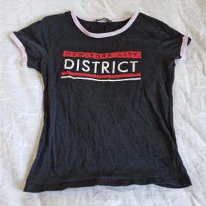 Svart t-shirt med tryck från FB Sister - Säljer en svart t-shirt från FB Sister med texten 'New York City District' i vitt och rött på framsidan. T-shirten har korta ärmar och en ljusrosa kant vid halsen och ärmsluten. Perfekt för en casual look!