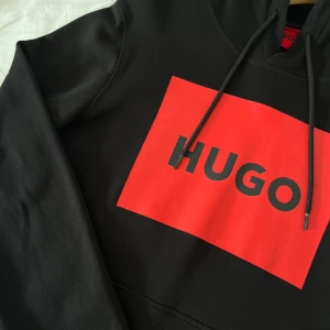  Hugo Boss Hoodie - Säljer en snygg svart hoodie från Hugo Boss med en stor röd logga på framsidan. Den är perfekt för både vardag och chill. Hoodien har en klassisk passform med långa ärmar och en praktisk ficka framtill. Priset går och diskutera 