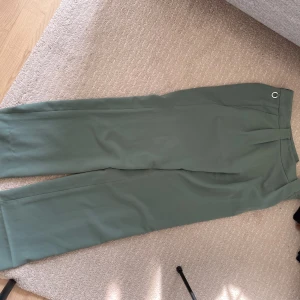 Gröna byxor från Stockh LM - Säljer ett par stilrena gröna byxor från Stockhlm. De har en snygg ringdetalj i midjan och är gjorda i ett bekvämt material. Perfekta för både vardag och fest!