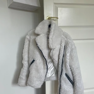 Vit teddyjacka från NA-KD - Supermysig vit teddyjacka från NA-KD som är perfekt för kyliga dagar. Jackan har en snygg asymmetrisk dragkedja och två fickor med dragkedjor. Insidan är fodrad med ett glansigt material som känns lyxigt mot huden. Den är både stilren och varm, vilket gör den idealisk för både höst och vinter.